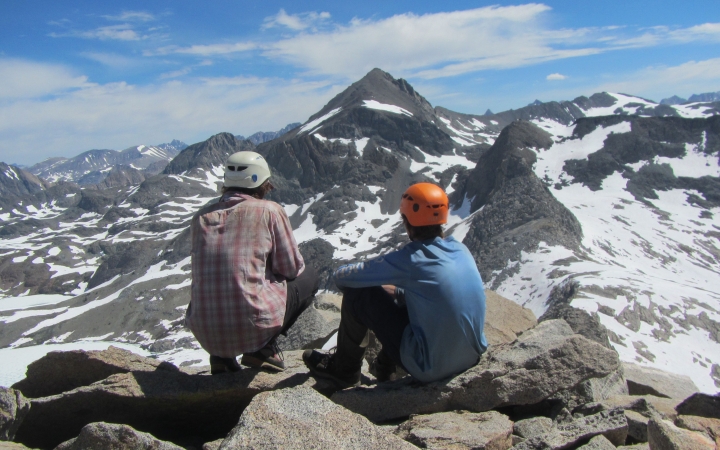 two people wearing helmets face away from the camera, looking at a vast and snowy mountainous landscape